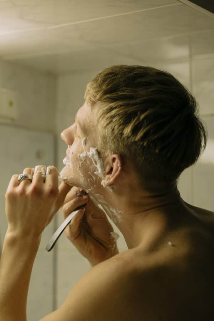 Shirtless man with blond hair shaving using a straight razor, focusing on self-care.